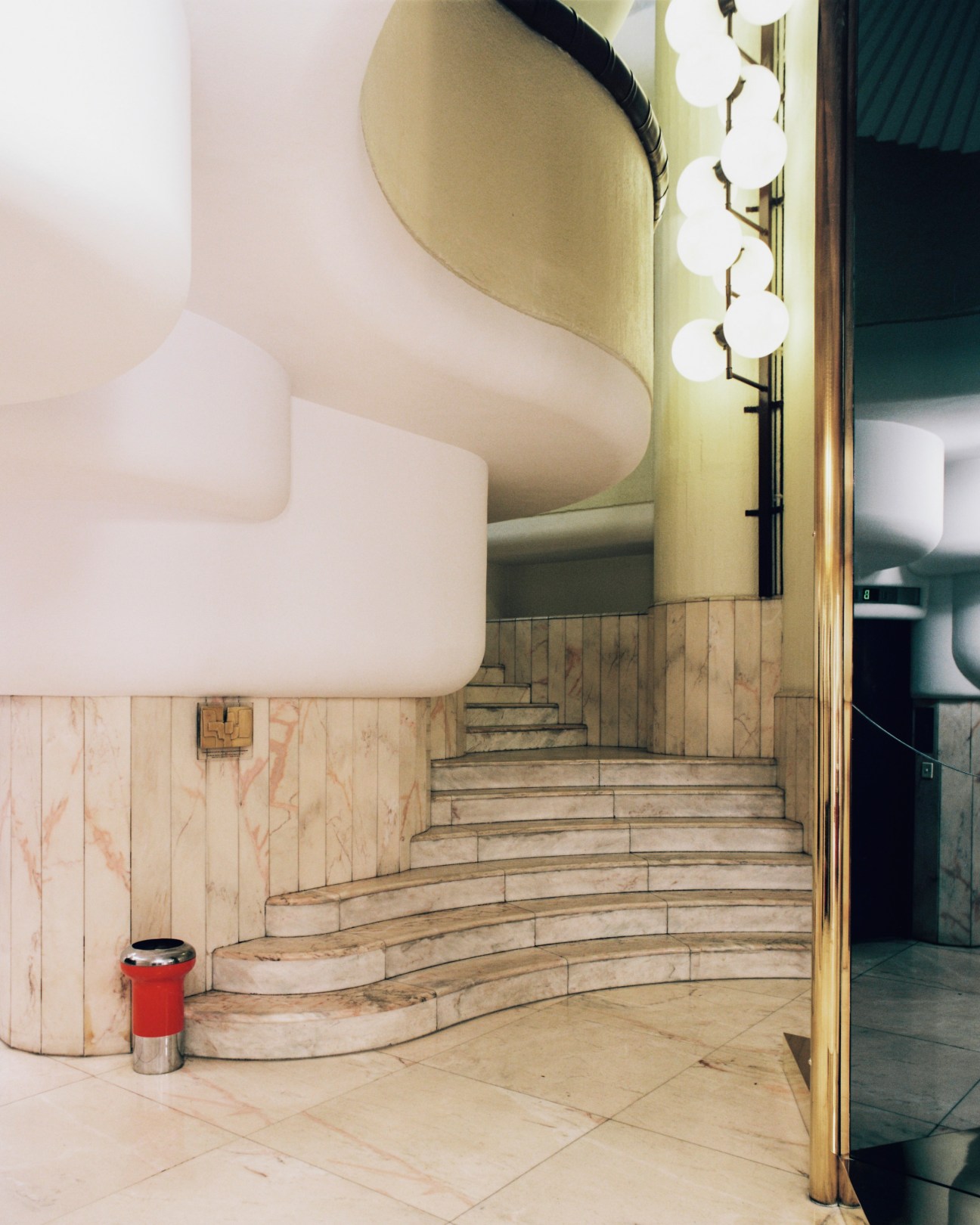 Base of central spiral staircase with curved white walls, pale marble flooring, and red cylindrical waste bin.