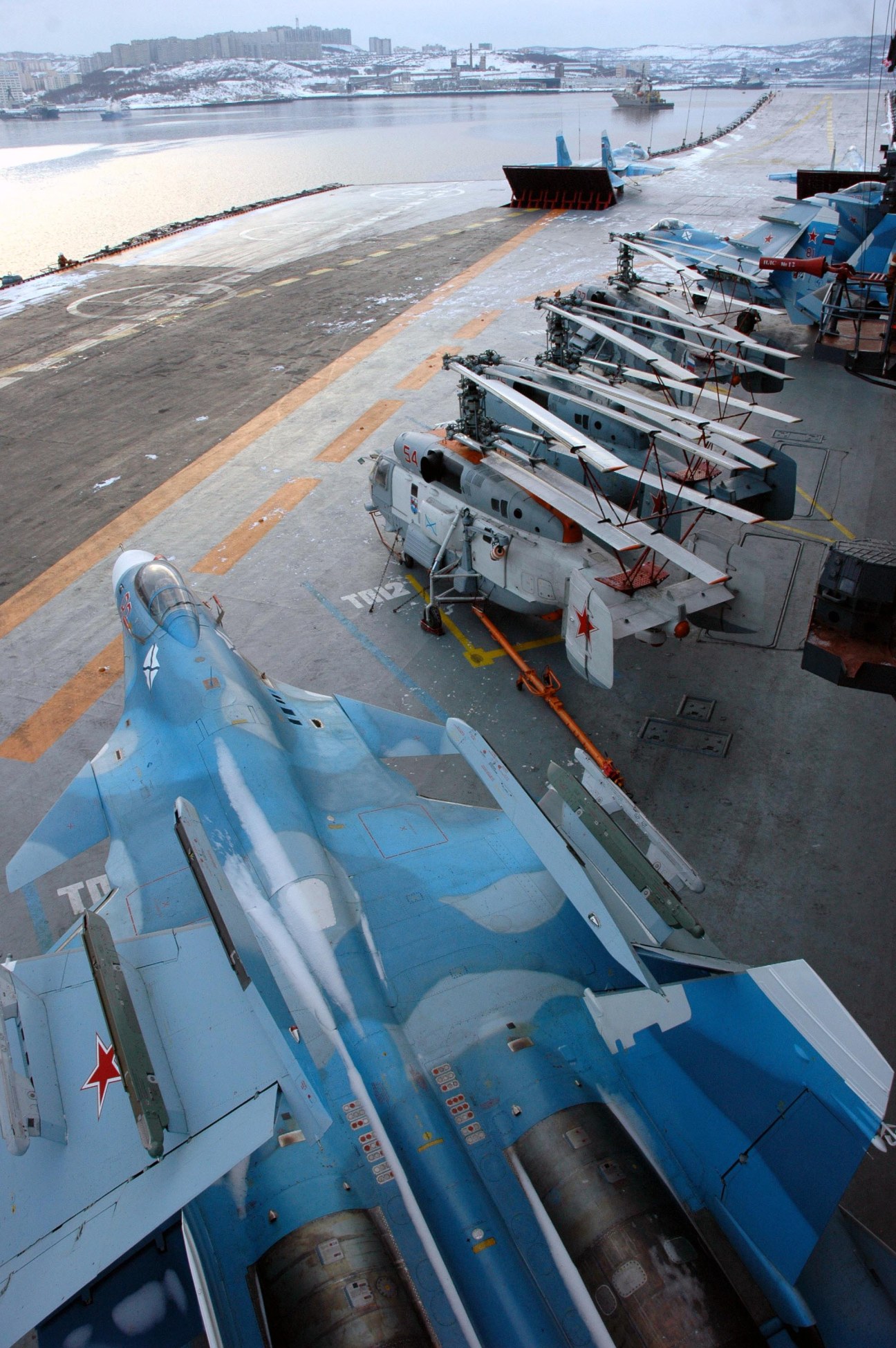 Russian fighter jets lined up on a snowy airfield with naval vessels visible in the background.