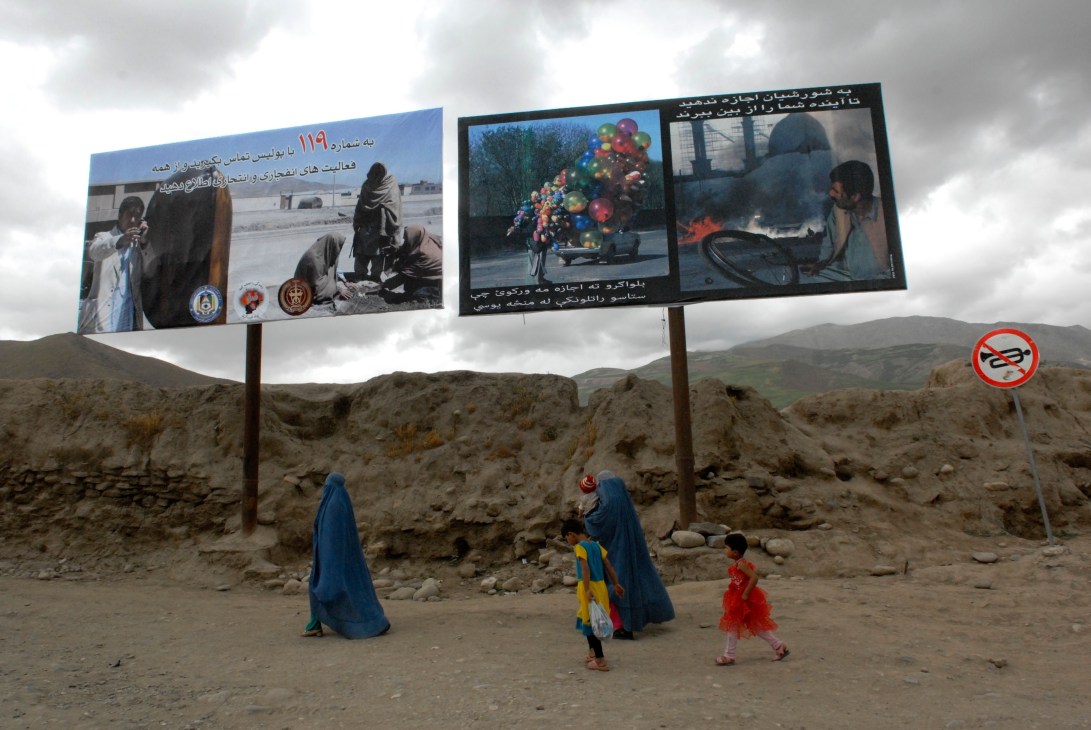 Afghan women and children walk past government anti-terrorism billboards in a rural mountainous area.
