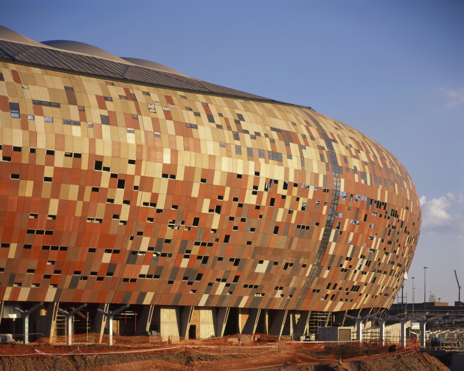 A large modern stadium with a distinctive brick and metal facade featuring a checkered pattern in orange, yellow, and tan tones.