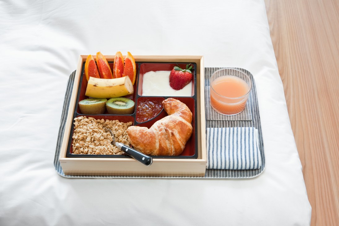 Breakfast bento box with croissant, fruit, yogurt, granola, juice and pastries on white bedding.