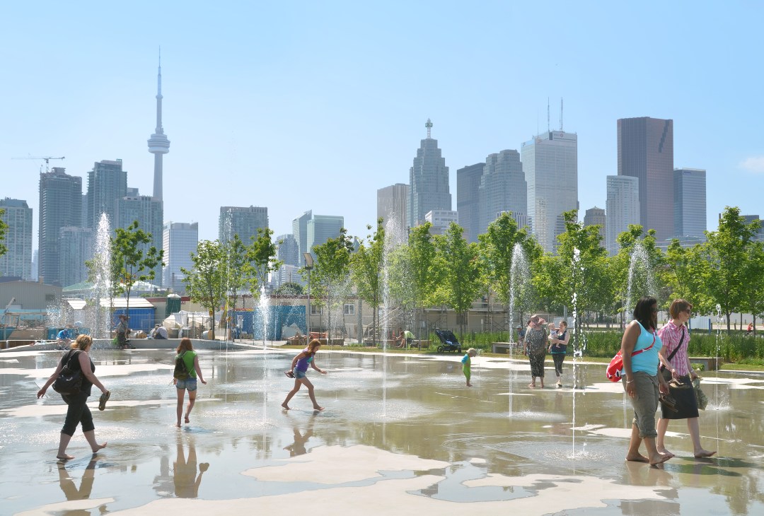 People play in fountains at a Toronto waterfront park with city skyline and CN Tower in background.