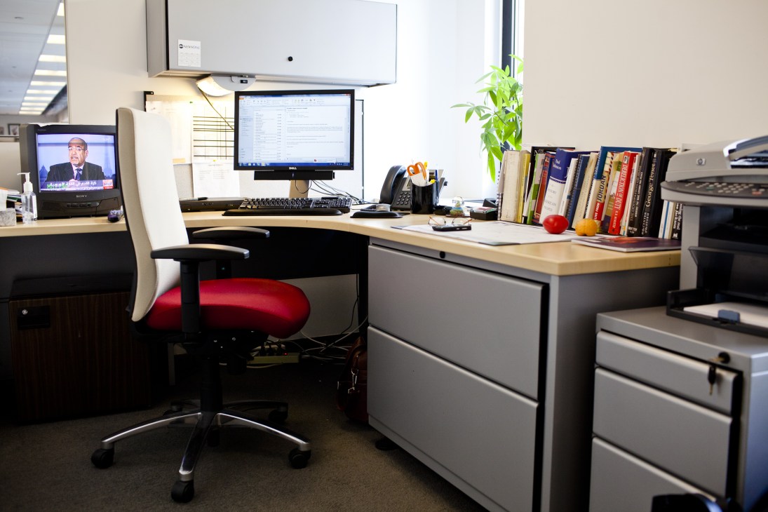 A modern office desk with computer monitors, books on politics, and a red office chair in a media bureau workspace.