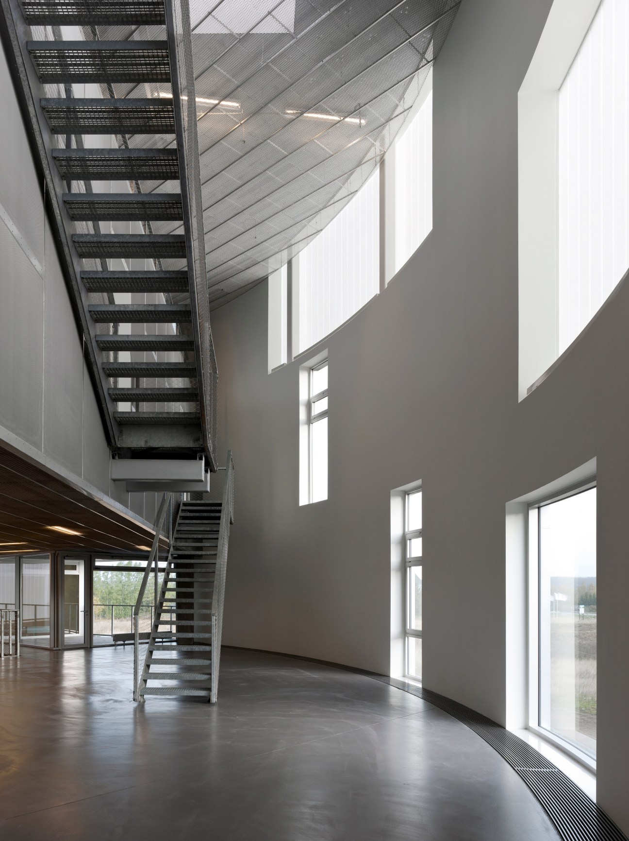 Angular industrial interior with metal staircase, corrugated glass ceiling, and white walls featuring rectangular windows.