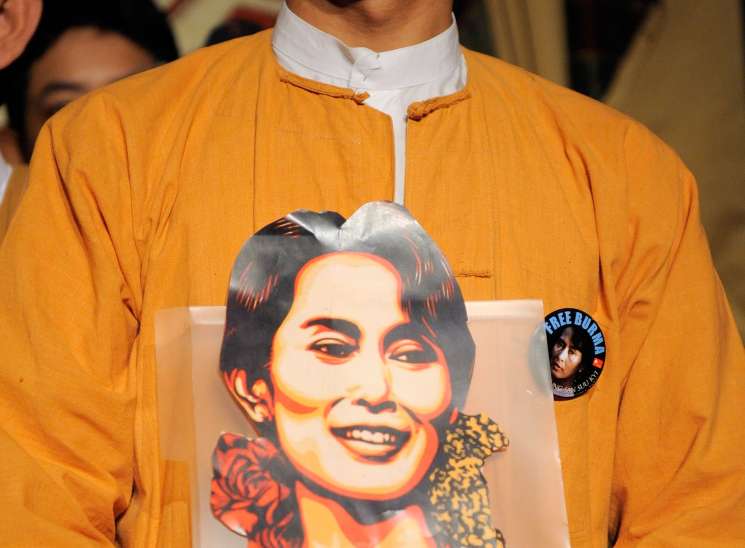 Man in orange shirt holding poster with portrait of Aung San Suu Kyi featuring political imagery and text.