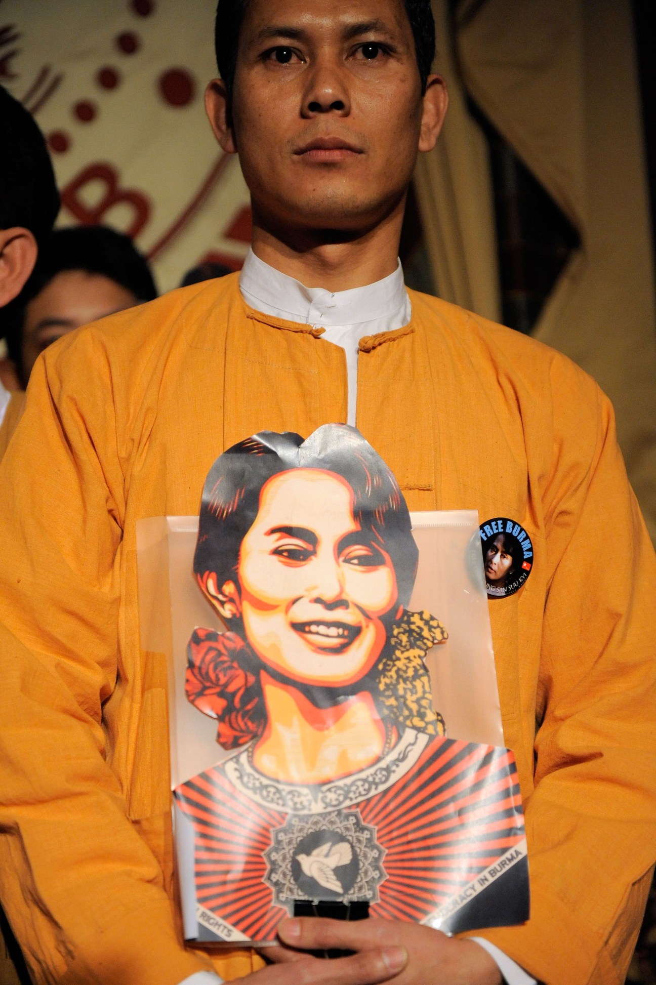 Man in orange shirt holding poster with portrait of Aung San Suu Kyi featuring political imagery and text.