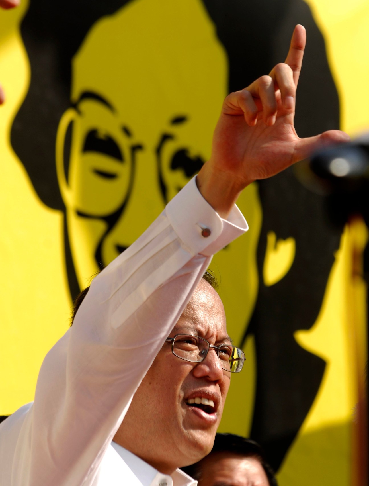 Man in glasses and white shirt making a peace sign with yellow propaganda posters in background.