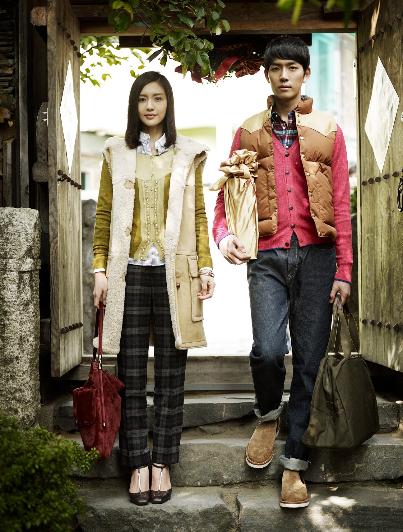 A young woman and man stand in a traditional Korean doorway wearing designer autumn clothing and holding bags.