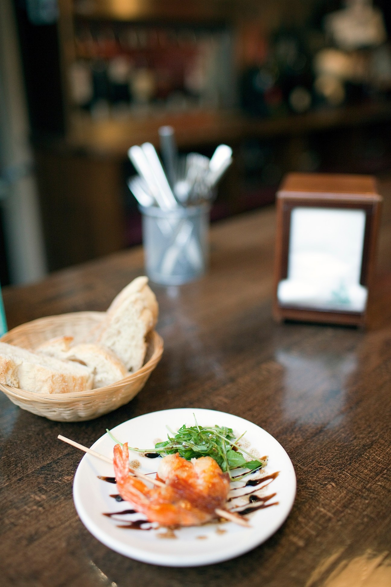 A plated pintxo with shrimp and microgreens on a white plate at a restaurant table with bread basket and napkins.