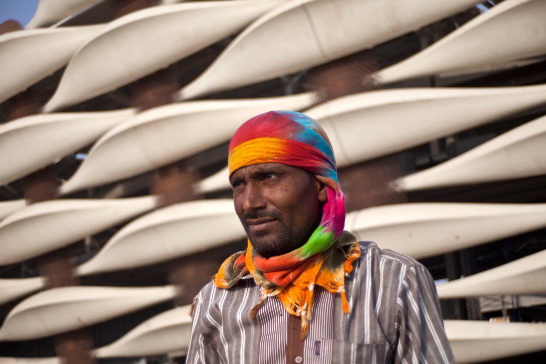 Indian worker Mahaveer wearing a colorful headscarf in front of large curved pipes at Basra Sports City construction site.