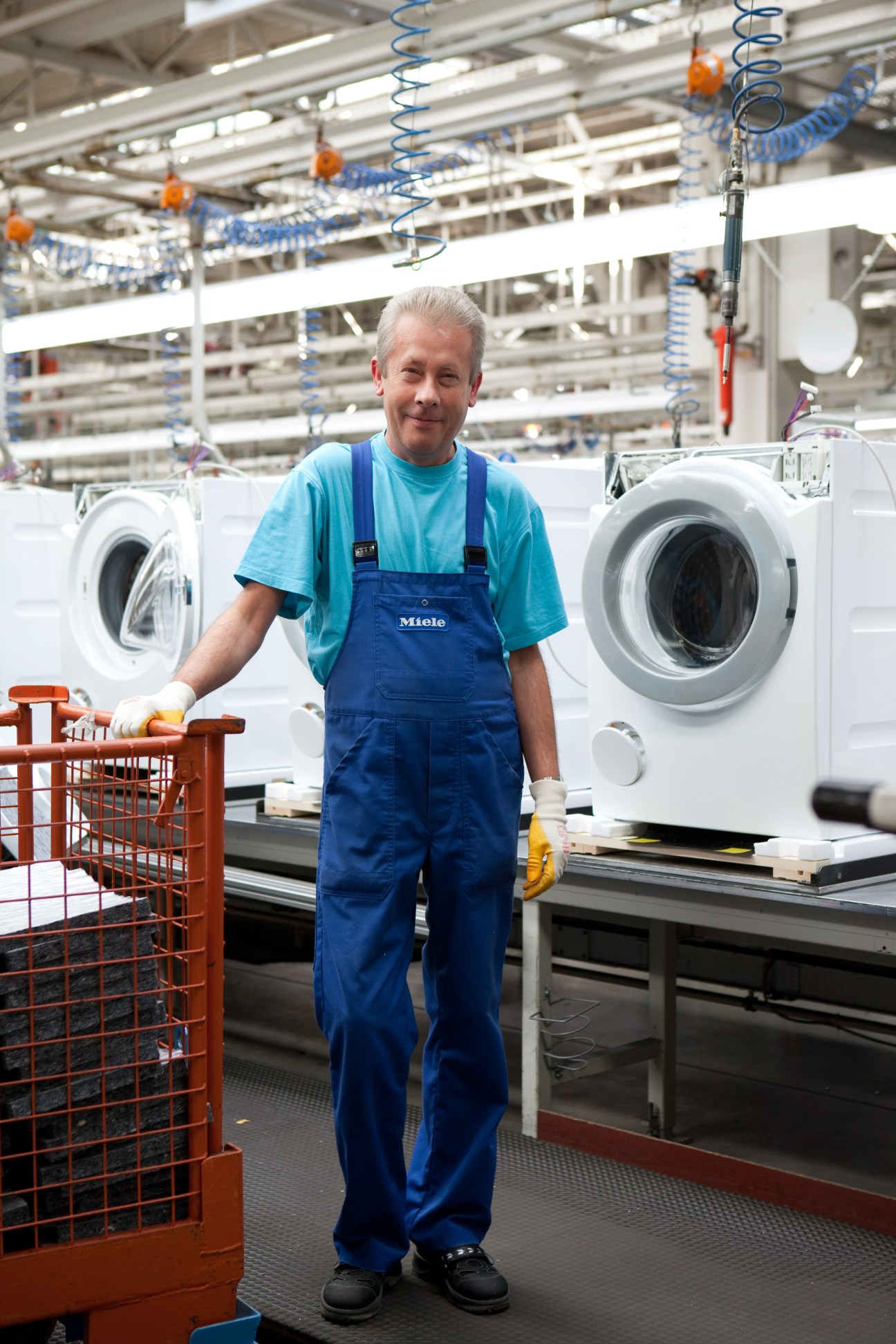 A Miele factory worker in blue overalls stands at a washing machine assembly station.