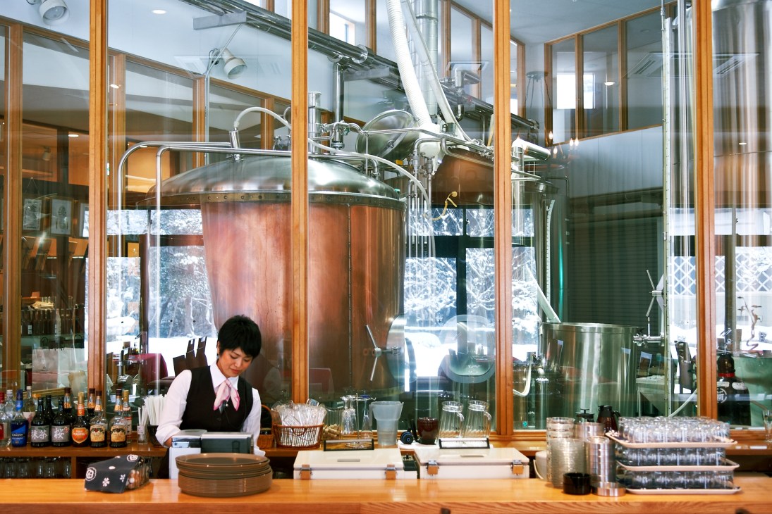 A bartender works behind a counter at Swan Lake brewery's restaurant with large copper and stainless steel brewing kettles visible through windows behind.