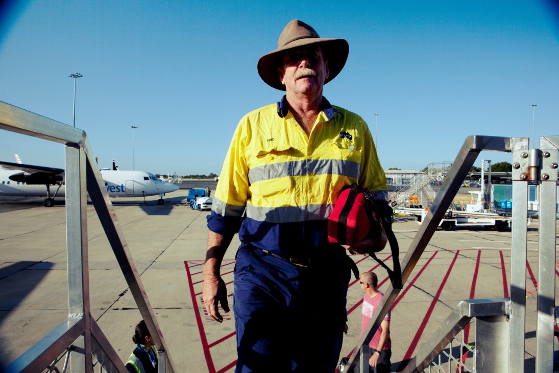 A fly-in fly-out worker in yellow and blue high-visibility work wear and cowboy hat boards an aircraft at Perth Airport.