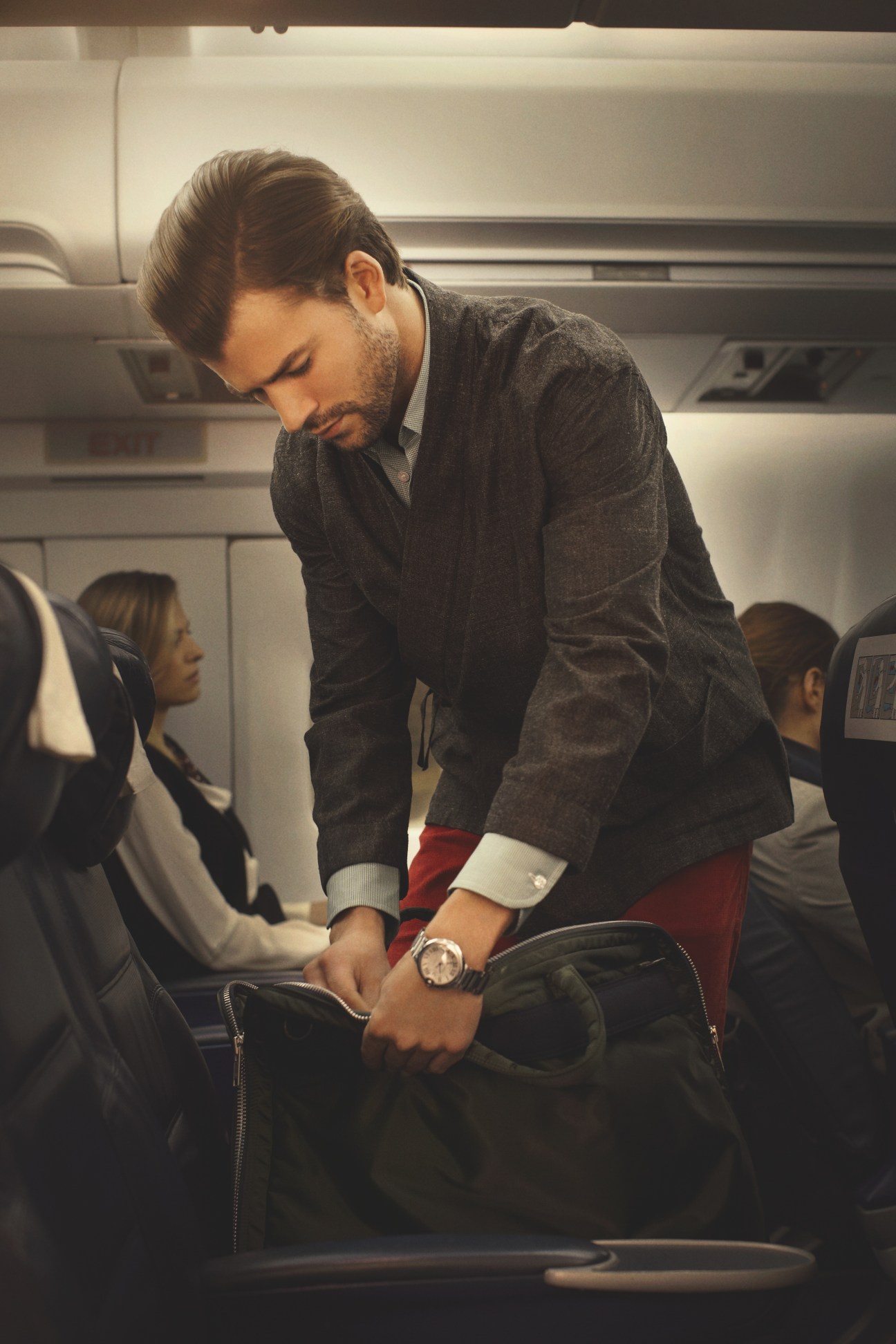 Man in airplane cabin wearing Louis Vuitton jacket and Cartier watch, adjusting his bag in the overhead compartment.