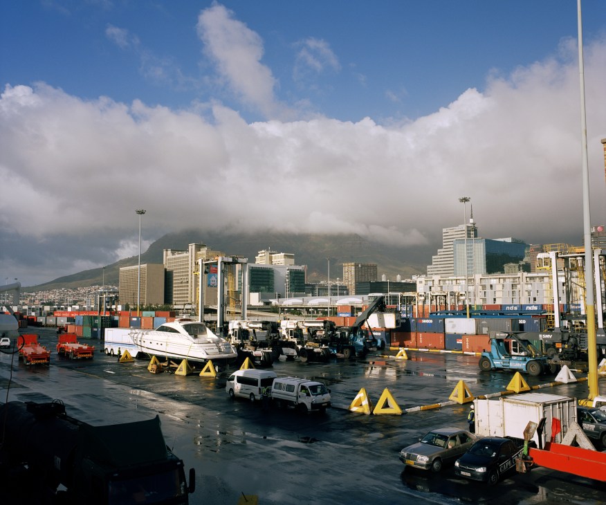 Cape Town shipping port with Table Mountain in background, showing cargo containers and vehicles on wet dock.