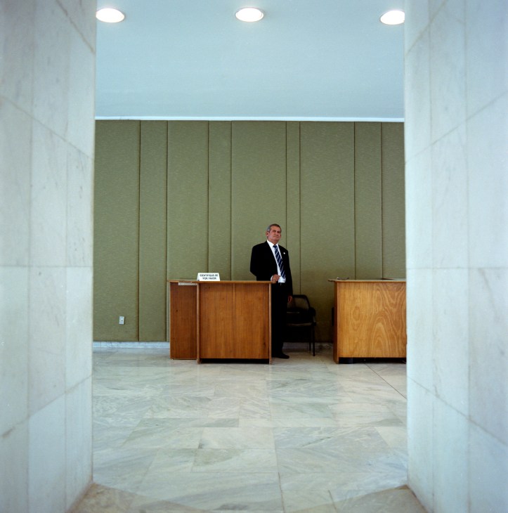 A security guard in a black suit stands in a minimalist corridor with white marble walls and light wood reception desks.