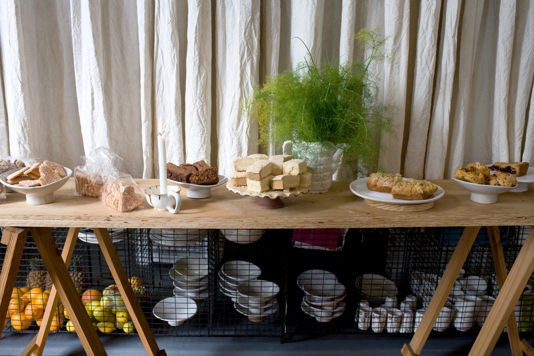 Wooden table display at Birds Boutique Café with baked goods, cheese, and fresh herbs against cream curtains.
