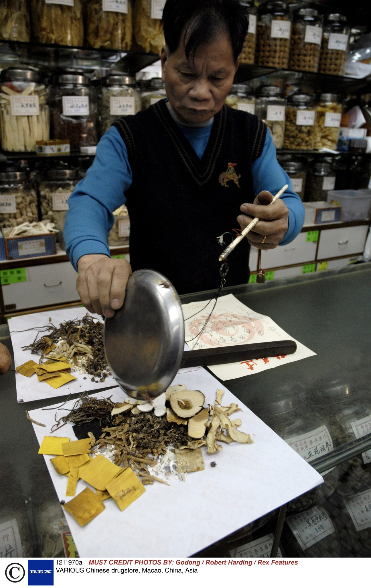 Woman in blue shirt weighing medicinal herbs in a traditional Chinese drugstore filled with labeled jars.