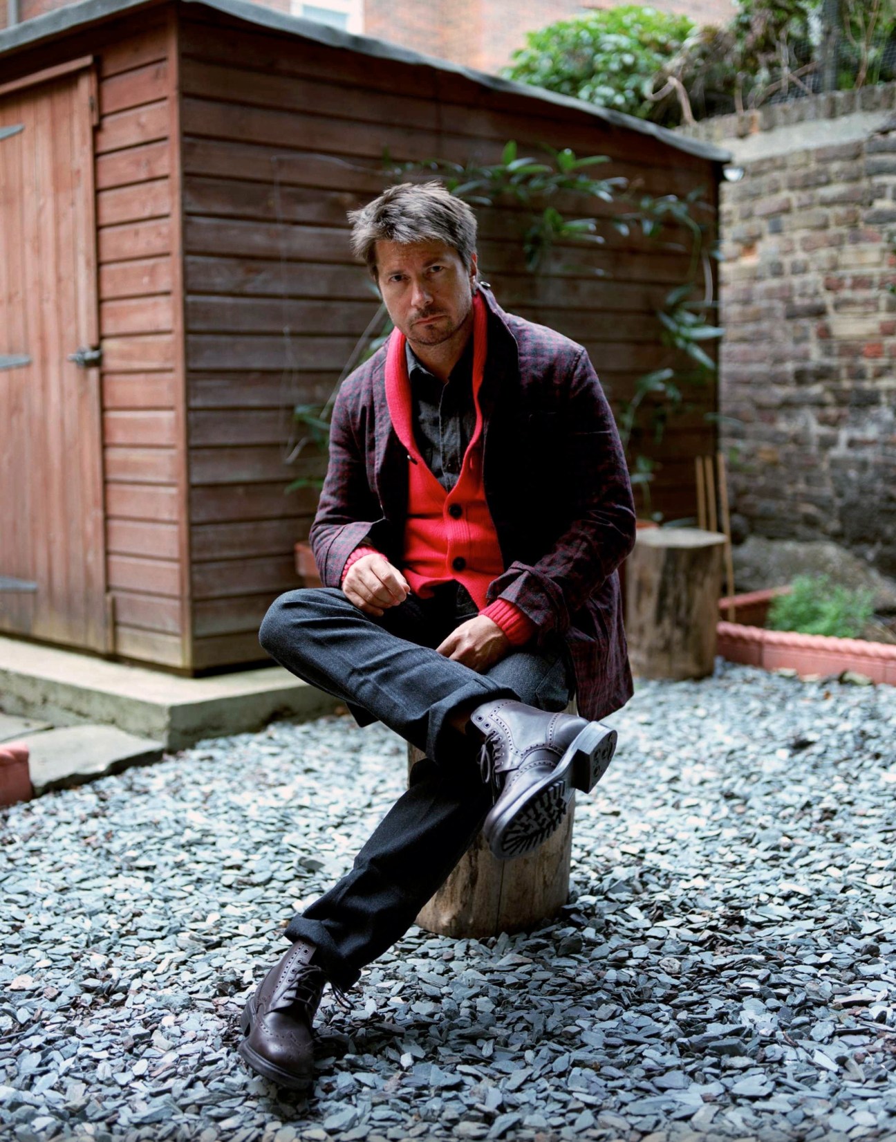 Man in burgundy cardigan and red scarf sitting outdoors against wooden shed backdrop.