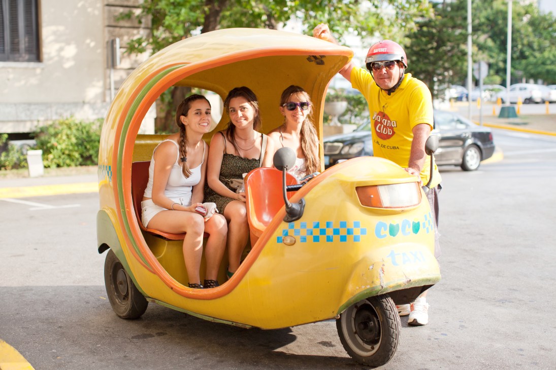 Tourists riding in a colorful yellow and orange vintage Cuban taxi with a driver in a yellow shirt and safety helmet.