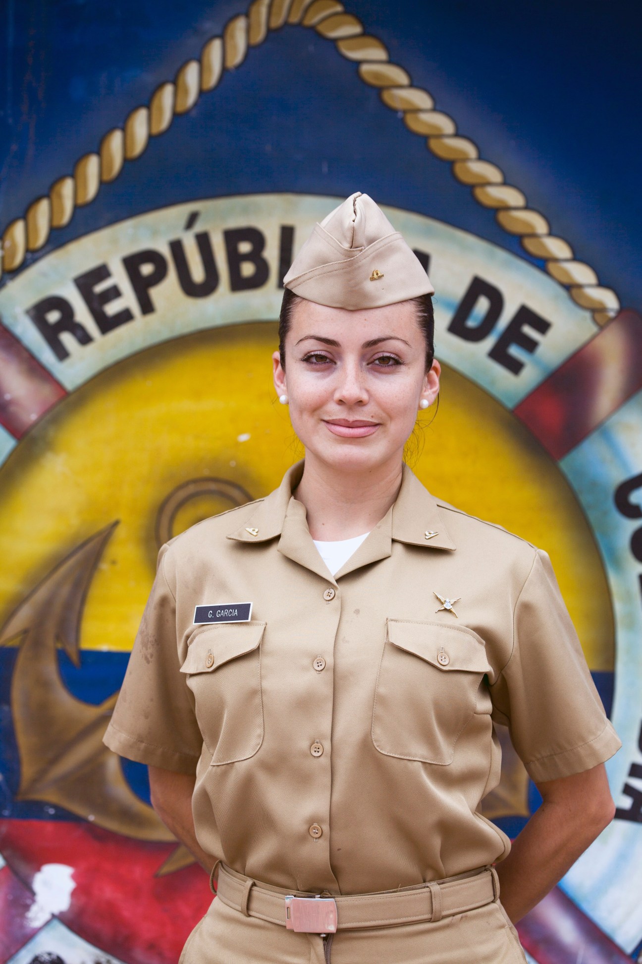 Naval officer Gloria García in tan uniform and white cap standing in front of Colombian flag seal.