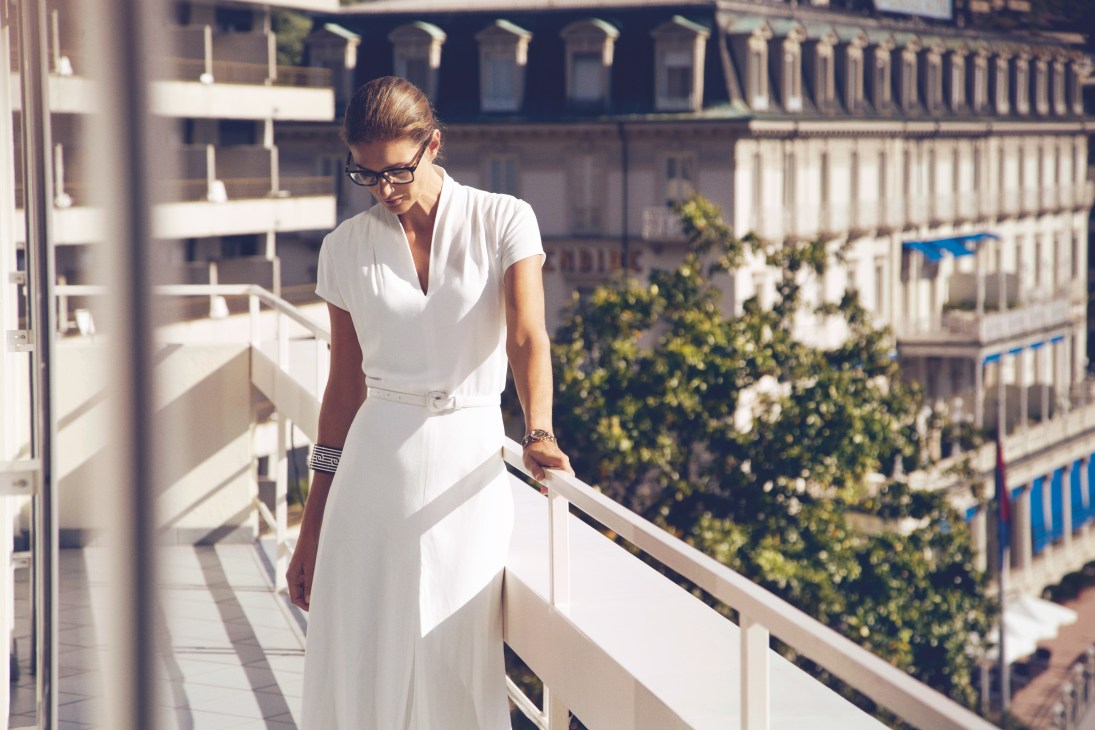 Woman in white Ralph Lauren dress and Prada glasses on a balcony overlooking Lugano buildings.
