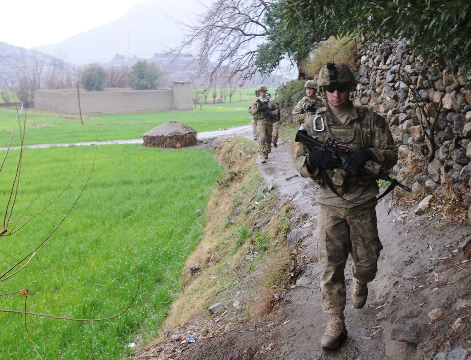 US soldiers in MultiCam camouflage uniforms patrol through mountainous Afghan terrain with stone walls and green fields.