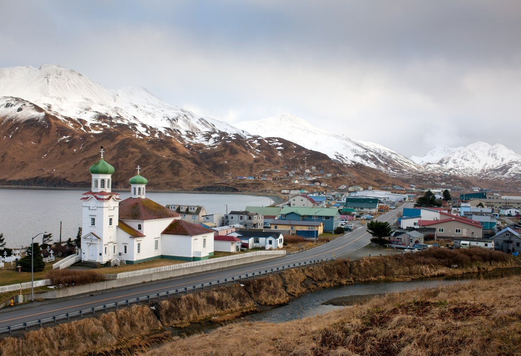 Snow-capped volcano overlooks Unalaska with Russian Orthodox church featuring green domes in foreground.