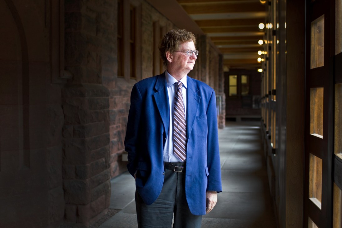 John Kirton stands in a brick corridor at the University of Toronto wearing a blue blazer and striped tie.