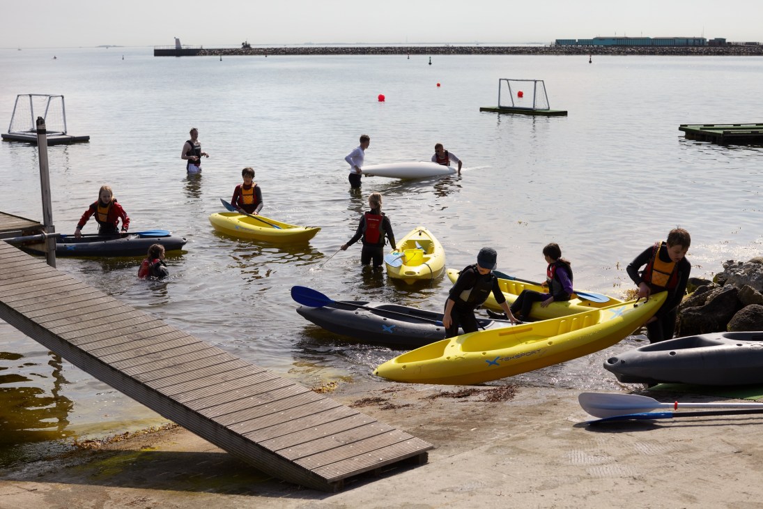 Children and adults with kayaks and water sports equipment in a shallow bay with a wooden dock.
