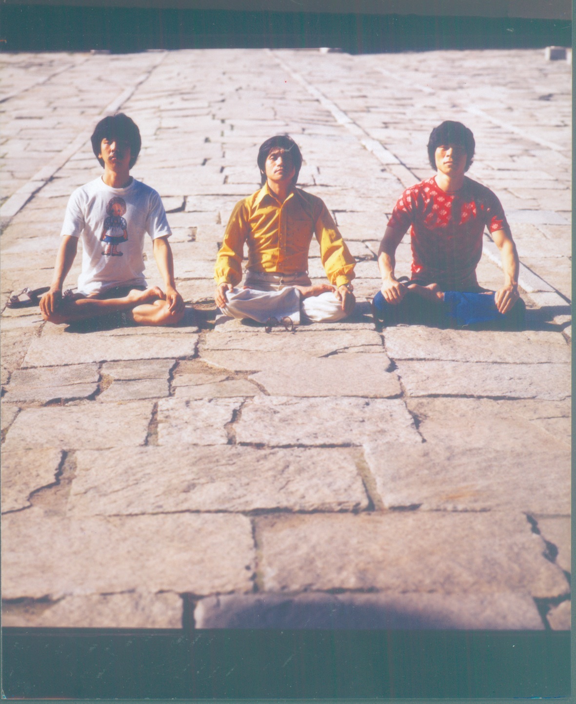 Three men with dark hair sitting cross-legged on stone pavement, wearing colorful 1970s-style shirts in white, yellow, and red.