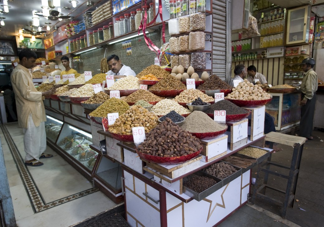 A spice market stall at Khari Baoli displays numerous varieties of dried spices, lentils, and legumes in mounds with price tags, with shelves of packaged goods and jars visible behind.
