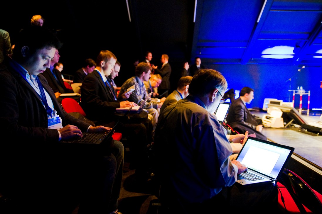 Attendees at a cyber security conference take notes on laptops in a darkly lit venue with blue lighting.