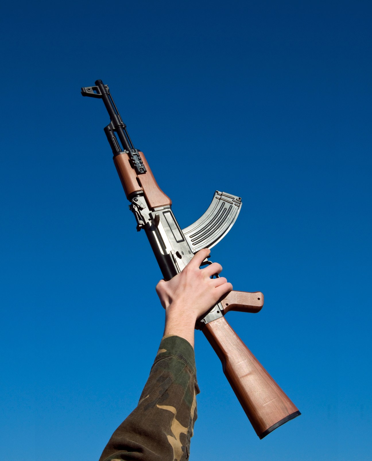 Hand holding an AK-47 assault rifle with wooden furniture against a blue sky.