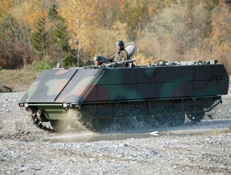 Military amphibious vehicle with armour plating carrying two soldiers in camouflage uniforms on gravel terrain.