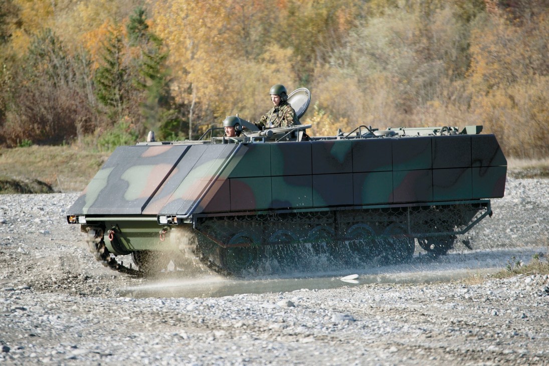 Military amphibious vehicle with armour plating carrying two soldiers in camouflage uniforms on gravel terrain.