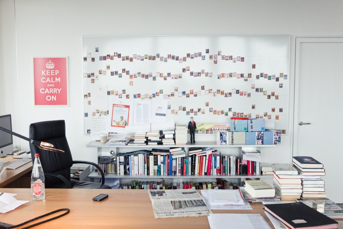 Office wall display with hundreds of small photographs arranged in rows above a desk with books and magazines.
