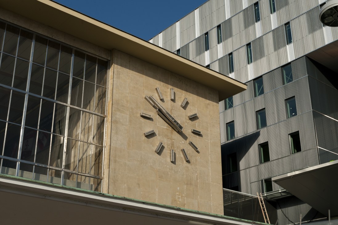 Modern railway station building facade with large clock and grid windows under blue sky.