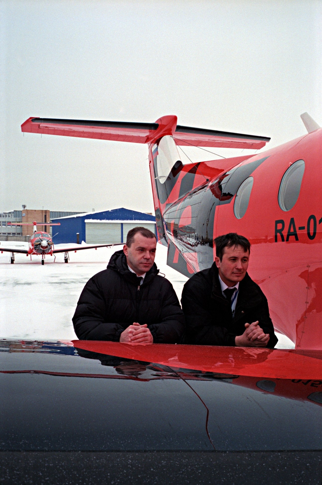 Two Dexter pilots Eduard Yevtyukhin and Sergei Tyurin stand beside a red Pilatus PC-12 aircraft on a snowy airport tarmac.
