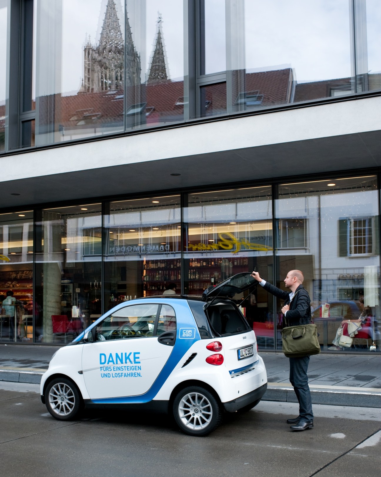 A man opens the door of a white and blue Smart car2go vehicle parked on a city street.