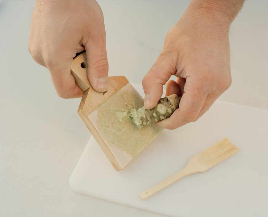 Hands grating fresh wasabi root on a wooden grater.