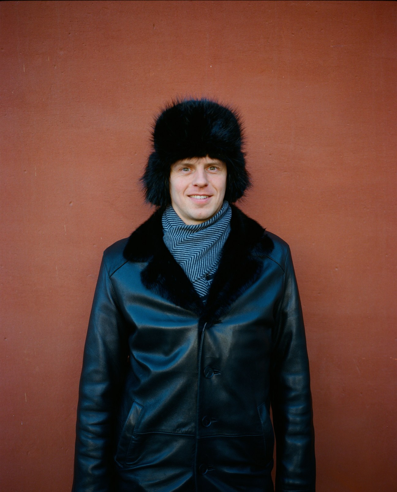 Man in black fur hat and blue leather jacket with striped scarf smiling against terracotta background.