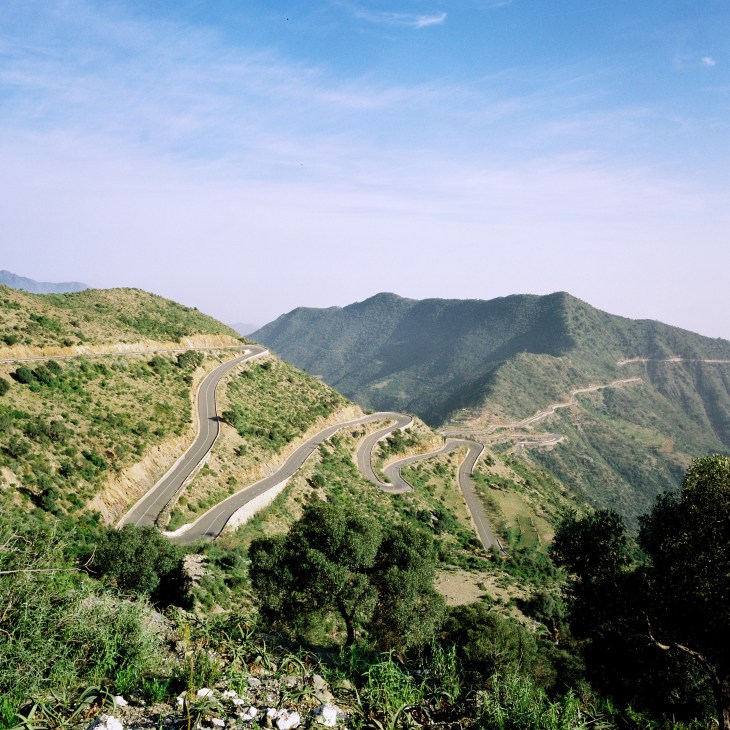 A winding paved road with sharp curves descends through green, mountainous terrain under clear blue skies.