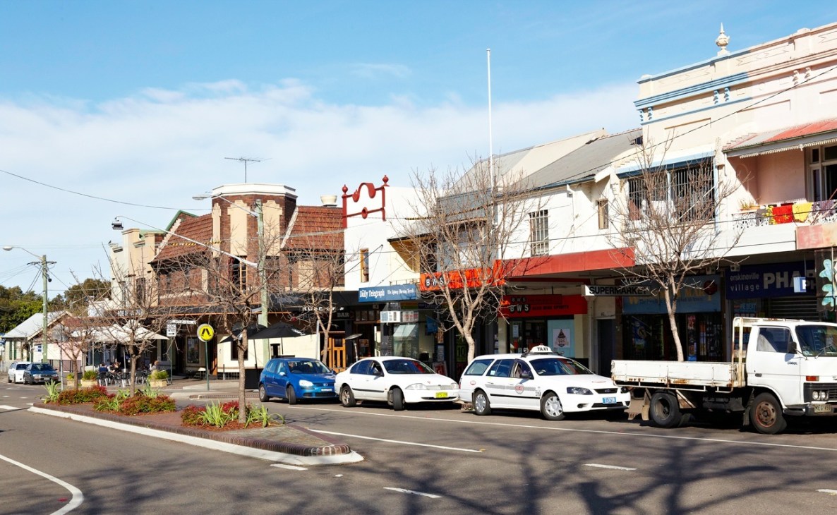 Erskineville Road street scene with Victorian-era shops, parked cars, and bare trees under blue sky.