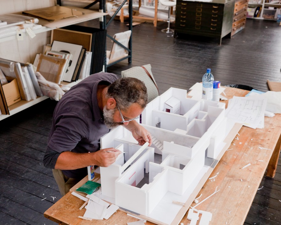 Man assembling a white architectural model at a wooden table in a studio workspace.