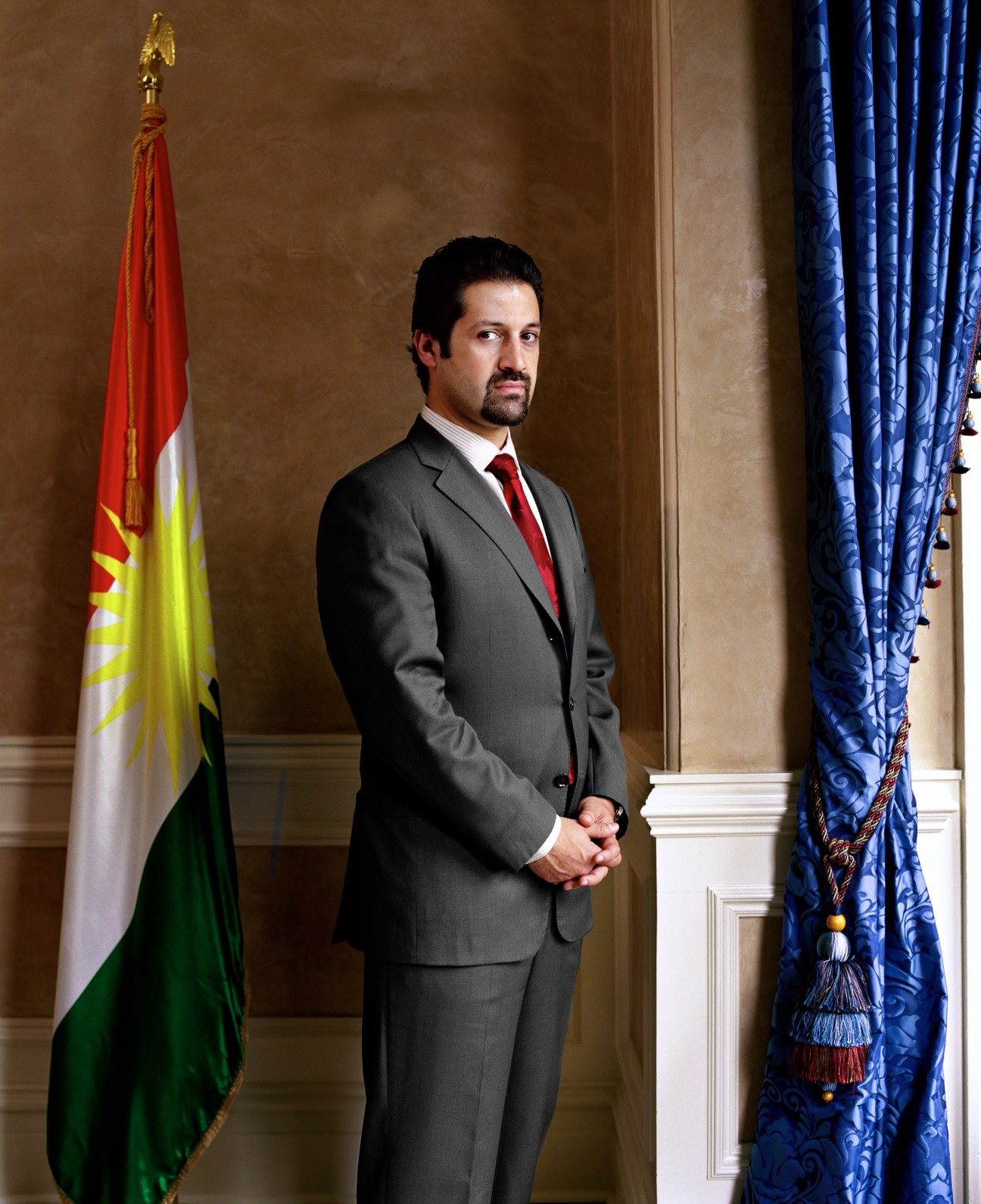 Qubad Talabani, a man in a gray suit and red tie, stands in a formal diplomatic setting with Kurdish flag and blue curtains.