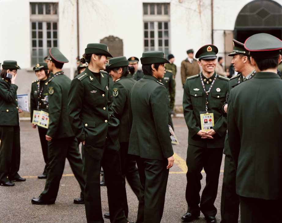 Chinese military athletes in green uniforms and caps stand conversing in a barracks courtyard during the Winter Military World Games.