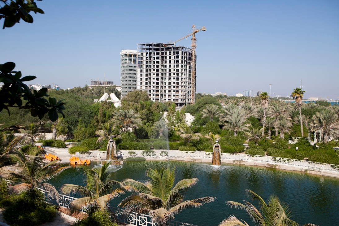 Unfinished high-rise building under construction on Kish island with artificial lake and palm trees in foreground.