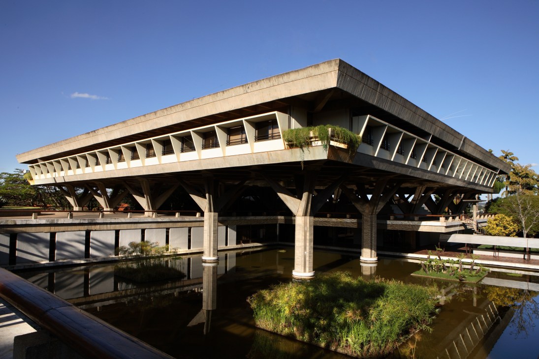 Modernist concrete building with horizontal lines and large windows reflected in surrounding water and tropical gardens.