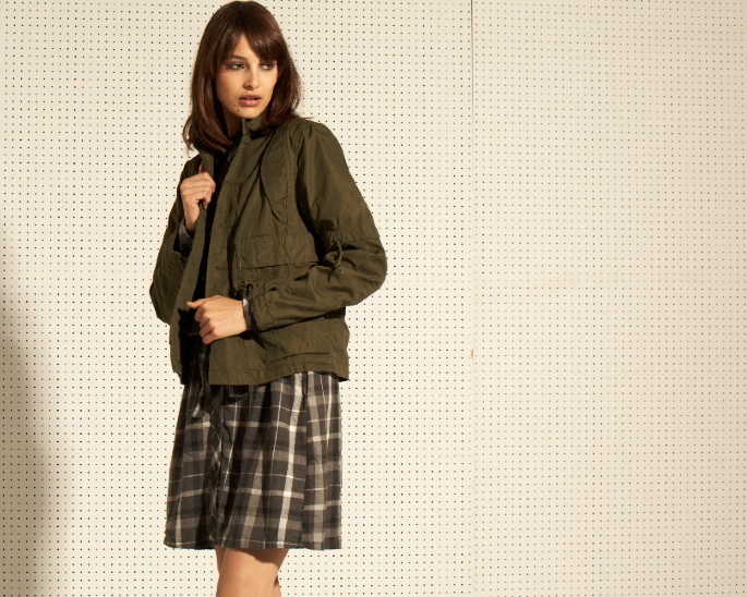 Woman wearing an olive green quilted jacket over a plaid skirt against a beige dotted backdrop.
